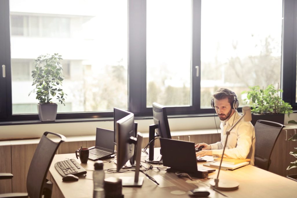 pexels photo 845451 A businessman sits at a desk using multiple computers and a headset in a well-lit modern office.